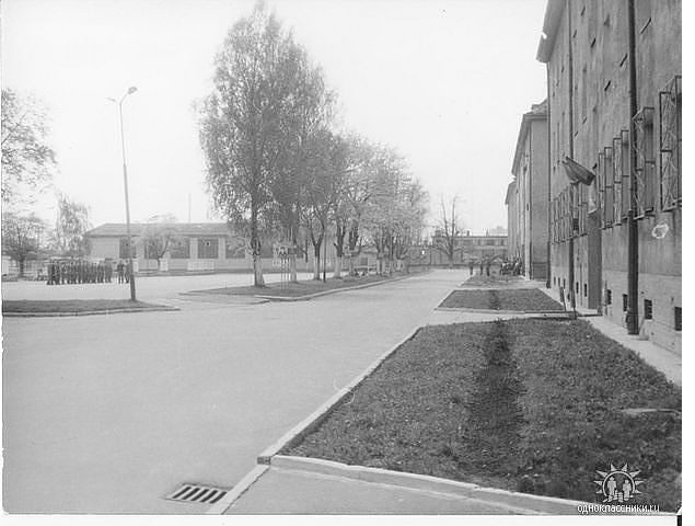 ROK 1982. Drzewa w wieku młodzieńczym . okratowane okna na parterze i grupka żołnierzy na placu alarmowym.